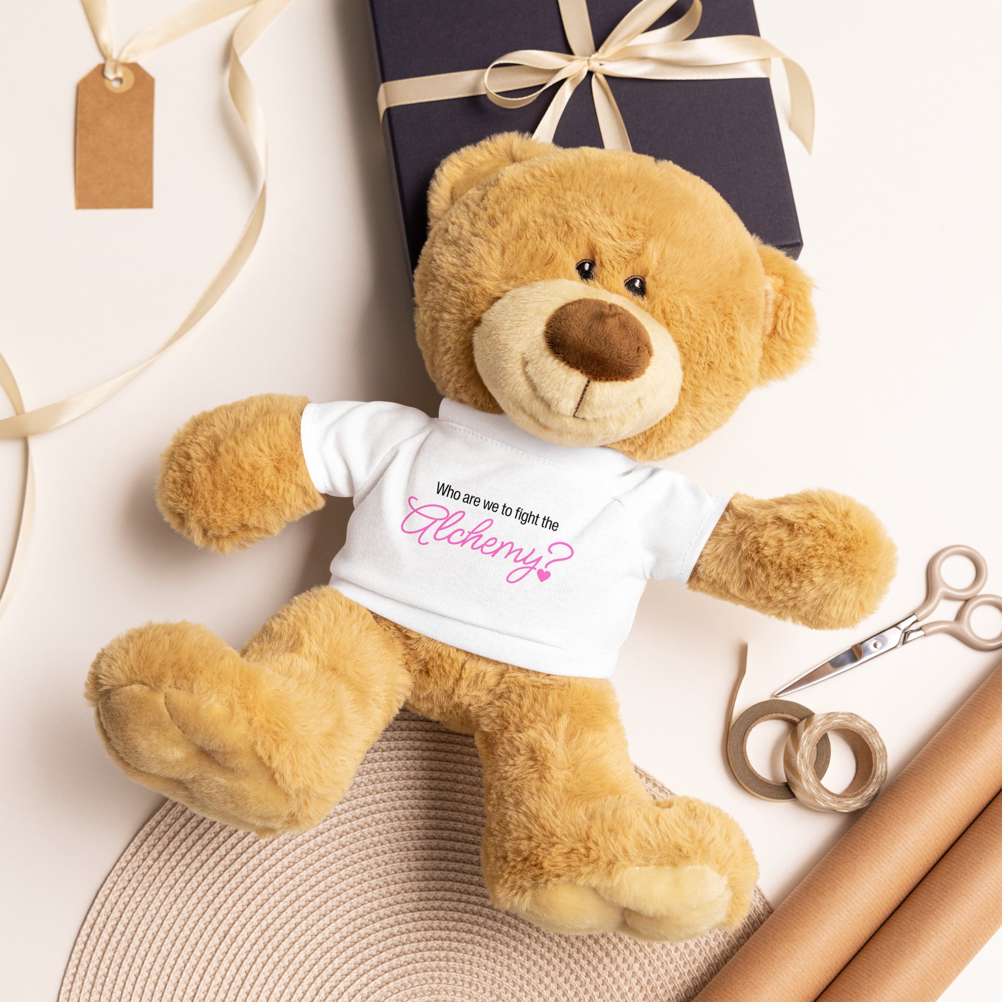 Teddy bear wearing a shirt with text, surrounded by gift wrapping materials on a light background